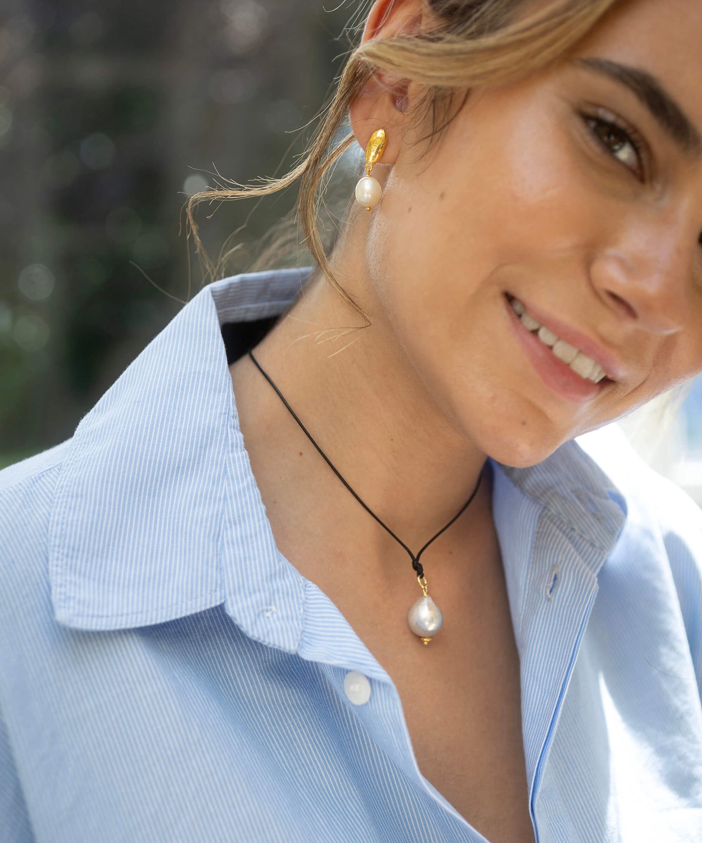 Woman wearing a pearl earring and necklace, smiling outdoors.