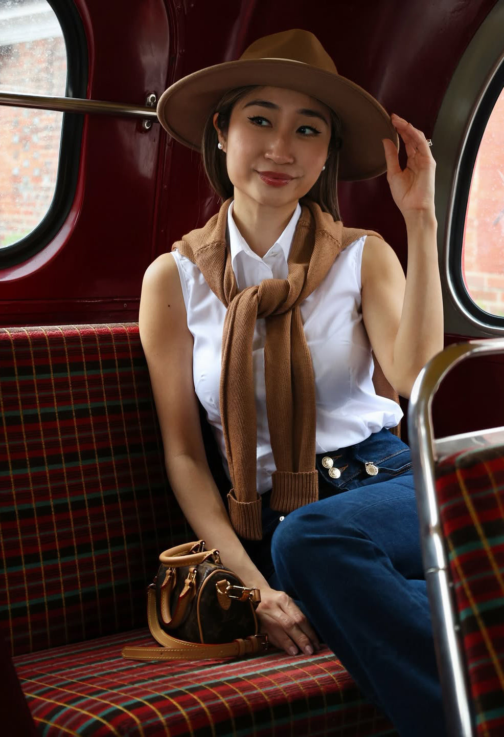 Woman sitting on a vintage bus wearing a brown hat and scarf, holding a handbag.